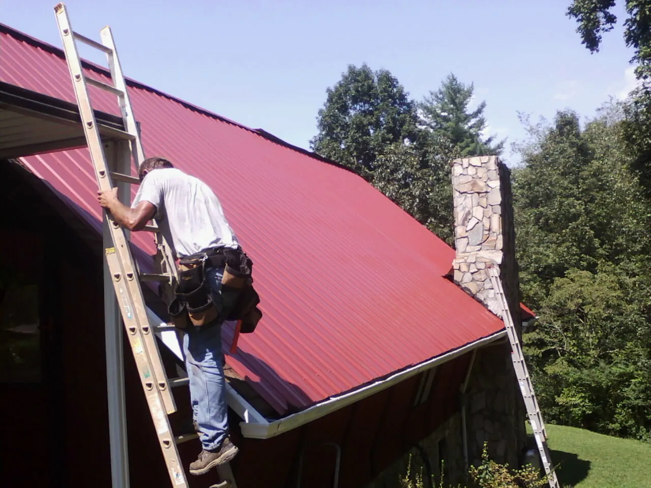 Roofer on a ladder beside a red metal roof and stone chimney — Soffit & Fascia Repair by Roofing Brothers in Highland Falls