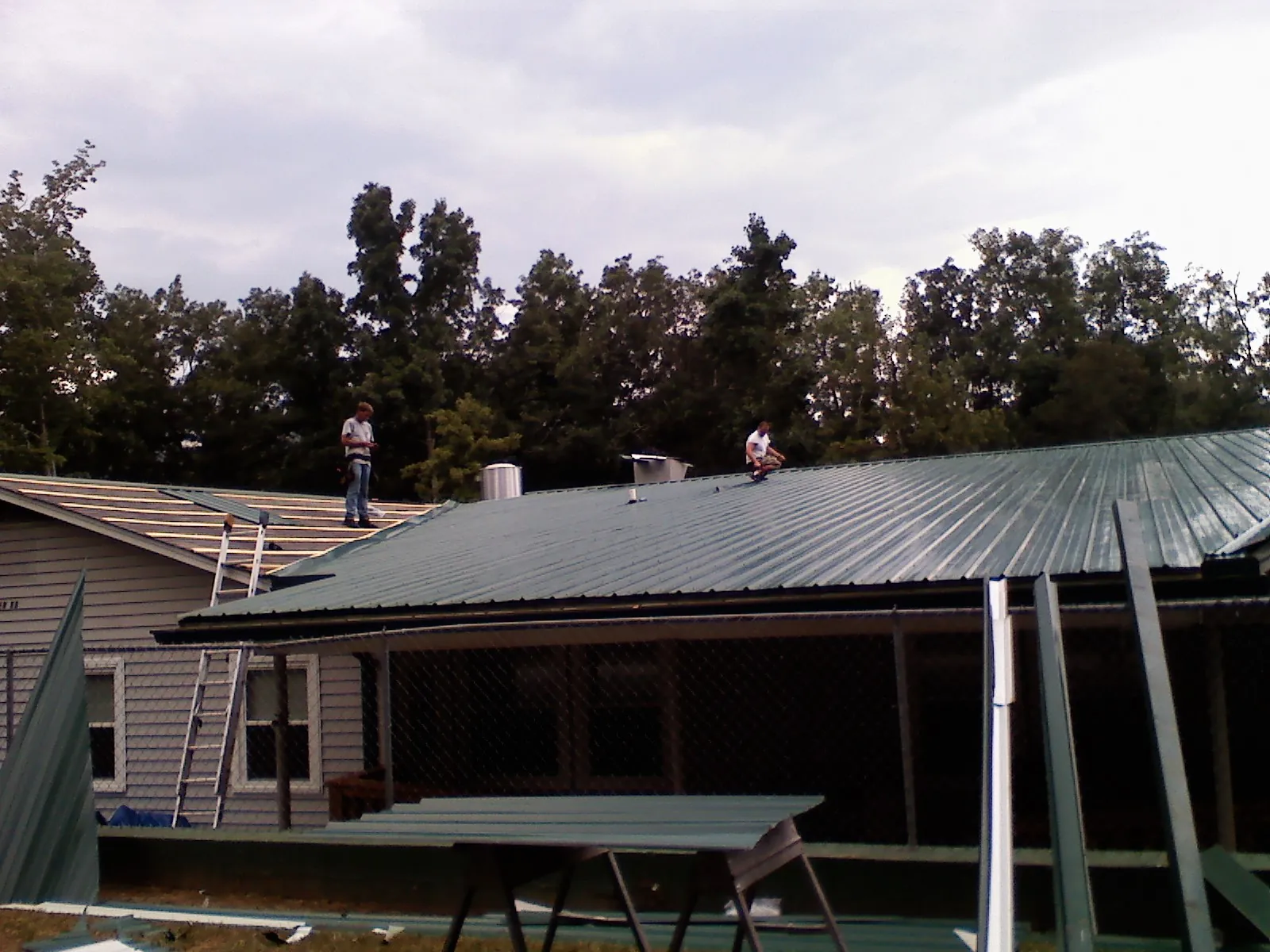 Two roofers installing green metal panels above a covered porch in Highland Falls